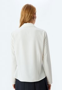 Blazer blanco de manga larga con cuello, diseño liso, textura suave y abertura en la parte posterior, combinado con una falda de rayas diplomáticas oscuras.