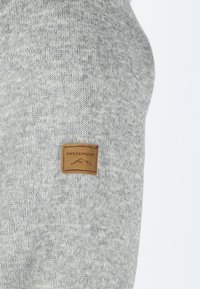 Tessuto in maglia grigia con un motivo strutturato. Presenta un piccolo etichetta in pelle marrone con la scritta "SWEDEMOUNT" vicino alla cucitura.