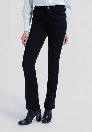 Žena u uskim crnim trapericama, crnim čizmama s petom i bijeloj košulji s gumbima, stoji nasuprot svijetlosivoj pozadini.