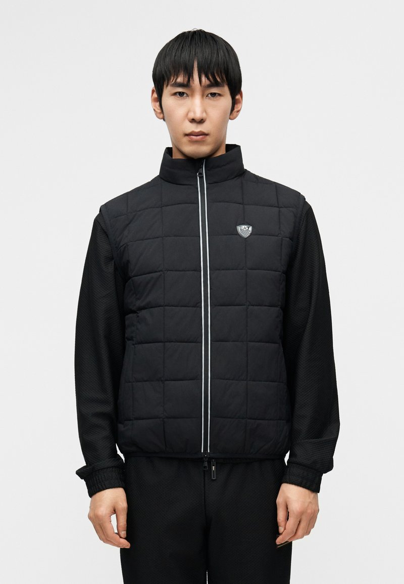 Μαύρο καπιτονέ γιλέκο με ψηλό γιακά, εμπρόσθιο φερμουάρ και λογότυπο, συνδυασμένο με υφασμάτινο μπλουζάκι με μακρύ μανίκι. Και τα δύο ρούχα έχουν μονόχρωμο σχέδιο.