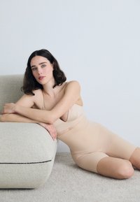 Beige shapewear met een aansluitend ontwerp, met een diepe V-hals en brede bandjes. Het model ligt op een genuanceerde beige bank.