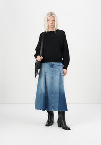 Schwarzer Pullover im Oversized-Schnitt, Jeansrock in Farbverlaufblau mit Paneeldesign und schwarze Knöchelstiefel. Das Model trägt eine Fransenhandtasche.