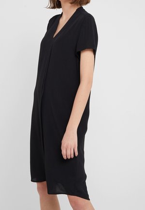 Kobieta ubrana w prostą czarną sukienkę do kolan z krótkimi rękawami i dekoltem w szpic, stojąca na jednolitym jasnym tle.