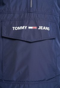Marinblå collegejacka i lättviktsmaterial, med en framficka som har "TOMMY JEANS"-logotype i vit text och rektangulär design.