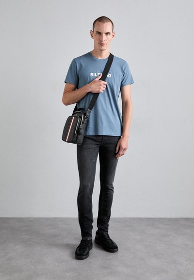 Geantă crossbody neagră cu o dunga albă și roșie, purtată peste un tricou albastru cu textul "SILENT", asociată cu blugi skinny închis și adidași negri.