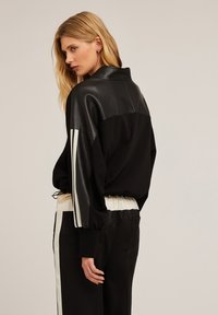 Schwarze Bomberjacke mit Lederoberteil, elastischem Bund und weißen Streifen an den Ärmeln. Strukturiert, mit lässigem Schnitt.