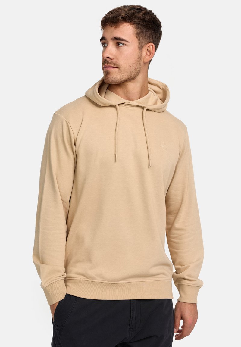 Young man with short brown hair wearing a beige hoodie and black pants, looking to his left against a plain light background.