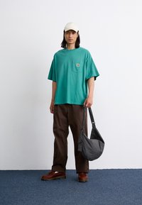 Tricou verde cu mânecă scurtă, cu buzunar și logo, pantaloni maro, pantofi maro, șosete albe și o geantă gri cu carouri, pe un fond simplu.