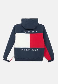 Μπλε ναυτικό μπουφάν τύπου windbreaker με κόκκινες και λευκές μπλοκ, κείμενο "TOMMY HILFIGER", κουκούλα, ελαστικά μανίκια και τελείωμα με αντιθετικές χρωματικές λεπτομέρειες.