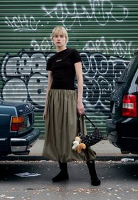 T-shirt preta justa, saia plissada verde azeitona, botas pretas até ao tornozelo e uma mala preta texturada com brinquedos de peluche. Fundo de graffiti urbano.