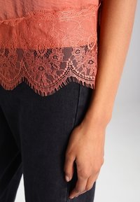 Koronkowy koralowy top z falbaniastym dołem, nałożony na czarny materiał, prezentujący skomplikowane detale koronkowe i gładką fakturę.