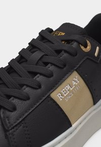 Μαύρο αθλητικό παπούτσι με παχιές μαύρες κορδόνια, μπεζ πλαϊνή επιφάνεια με το κείμενο "REPLAY SINCE 1981" και χρυσή λεπτομέρεια οπής κοντά στον αστράγαλο.