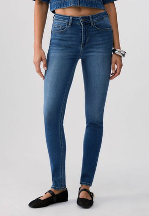 Frau trägt hochtaillierte blaue Skinny-Jeans, verkürzte Jeansjacke, schwarze Mary Jane Ballerinas und silberne Armbänder am linken Handgelenk, steht.