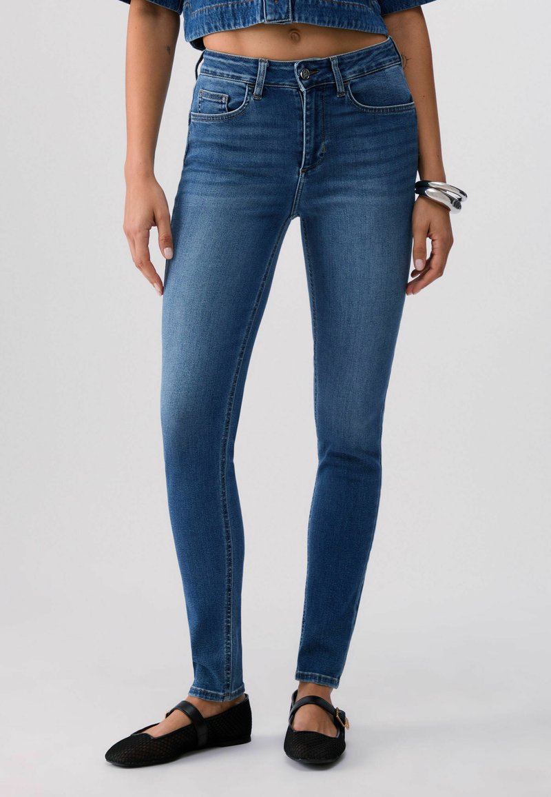 Frau trägt hochtaillierte blaue Skinny-Jeans, verkürzte Jeansjacke, schwarze Mary Jane Ballerinas und silberne Armbänder am linken Handgelenk, steht.