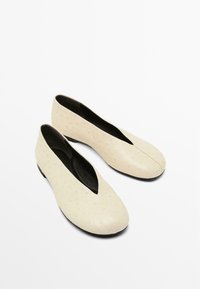 Scarpe slip-on beige con superficie testurizzata e un'apertura a forma di V. L'interno è nero e la suola è piatta e rotonda.