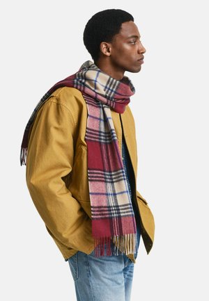 Junger Mann in senfgelber Jacke und blauen Jeans, der einen rot-, weiß- und blaukarierten Schal um den Hals gewickelt hat und nach rechts schaut.