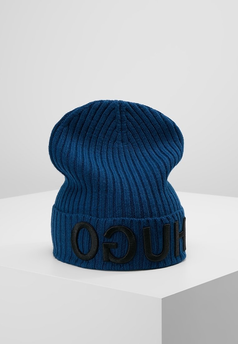 Bonnet bleu en tricot côtelé avec revers plié portant des lettres noires en relief formant le mot "HUGO", présenté sur une surface blanche.