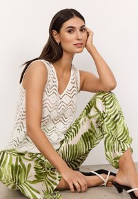 Weißes Zickzack-Spitzen-Top, grüne Hosen mit Blätter-Muster und schwarze Absatzsandalen. Model sitzt auf grauem Boden vor neutralem Hintergrund.