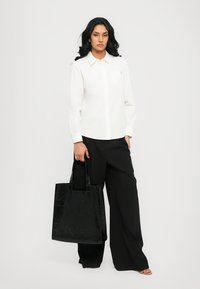 Cămașă albă cu butoni, pantaloni negri cu croi larg și o geantă mare neagră cu un textură netedă. Modelul stă pe un fundal neutr.