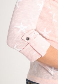 Ljusrosa långärmad skjorta med vita sjöstjärnemönster. Rullade ärmar fästa med knappdetalj. Mjuk, texturerad tyg.