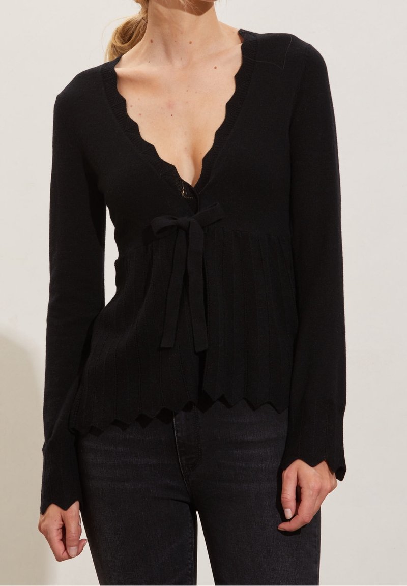 Femme portant un cardigan en maille noir à manches longues avec bords festonnés et détail noué, associé à un jean noir.