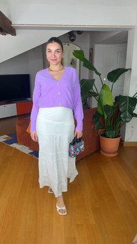 Frau in lila Strickjacke und weißem Rock, die eine Blumentasche hält und auf einem Holzboden neben einer großen grünen Pflanze im Innenraum steht.