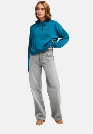 Junge Frau trägt einen türkisfarbenen Hoodie, hellgraue weite Jeans und beige Schuhe, steht mit einer Hand in der Tasche und schaut zur Seite.