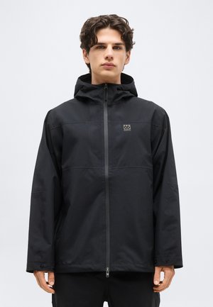 Junger Mann mit dunklem, lockigem Haar, der eine schwarze Kapuzenjacke mit Reißverschluss und dem "66 North"-Logo auf der Brust trägt, vor einem schlichten hellen Hintergrund steht.