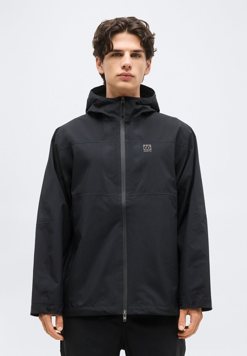 Jeune homme aux cheveux bouclés foncés portant une veste à capuche noire zippée avec le logo "66 North" sur la poitrine, debout devant un fond clair uni.