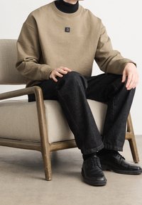 Persona seduta su una poltrona beige, indossa una felpa beige con logo, un dolcevita nero, jeans scuri e scarpe nere, con le mani appoggiate sulle ginocchia.