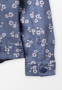 Tessuto di denim blu con piccolo motivo floreale bianco, cuciture a vista e un bottone blu scuro su un polsino di un indumento.