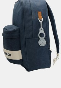Sac à dos en tissu bleu foncé avec une sangle beige et une poche avant, doté d'un porte-clés circulaire gris fixé à la tirette de la fermeture éclair latérale.