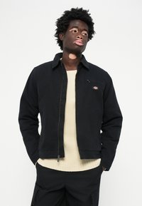 Chaqueta negra con cremallera y cuello, combinada con un suéter de punto color crema. Pantalones negros y cabello texturizado a la vista.