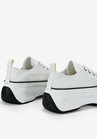 White canvas sneakers with black rubber soles, featuring rounded toes, lace-up front, and pull tabs at the back for easy wear.