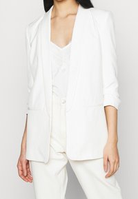 White blazer with a relaxed fit, notched lapels, and rolled sleeves. Front pockets and smooth texture, layered over a lace top.