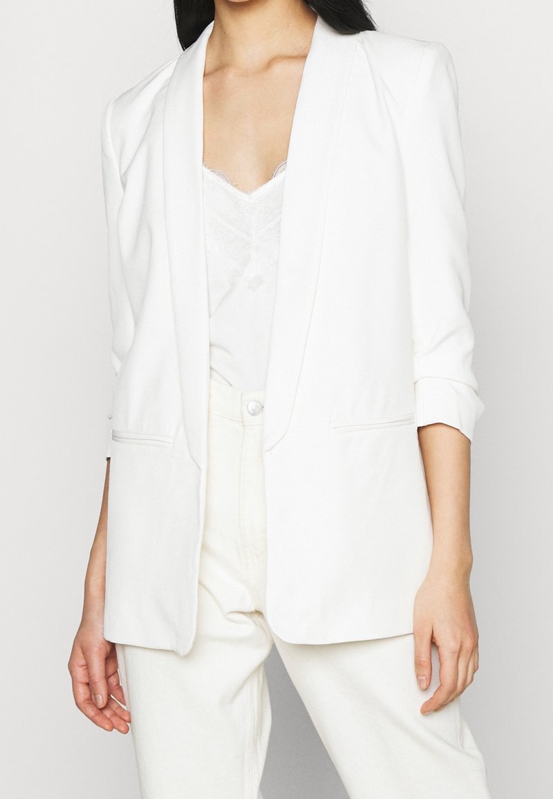 White blazer with a relaxed fit, notched lapels, and rolled sleeves. Front pockets and smooth texture, layered over a lace top.