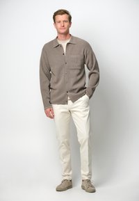 Brun ullskjorta med knappar och bröstficka, bärs över en ljusbeige t-shirt, tillsammans med krämfärgade byxor och beiga sneakers.