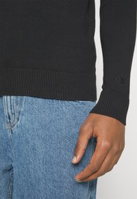 Maglione nero in maglia con orlo e polsini a costine. Presenta un logo ricamato sulla manica. Abbinato a jeans in denim azzurro chiaro.