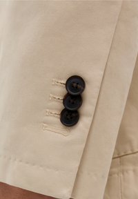 Tela de algodón beige con un corte ajustado, que presenta tres botones marrón oscuro en el puño y detalles de costura sutiles.