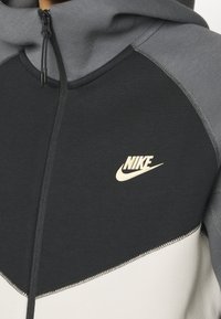 Nike mikina s kapucí na zip v černé, šedé a krémové barvě. Má klokánkovou kapsu, žebrované manžety a zlaté logo na hrudi. Hladká textura látky.