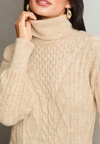 Maglione color crema a collo alto con motivi a treccia testurizzati e polsini a coste; tessuto morbido e caldo adatto per il clima più fresco.
