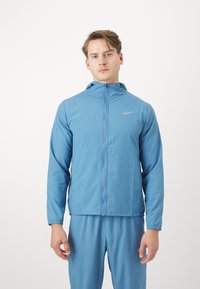 Lyseblå Nike jakke med fuld lynlås og justerbar hætte, udstyret med elastiske manchetter og logo på brystet. Bæres med matchende bukser.