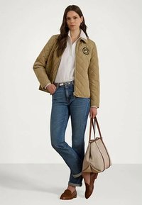 Femeie îmbrăcată cu o geacă bej matlasată, cămașă albă, blugi albaștri, mocasini maro, ținând o geantă bej cu mânere maro.