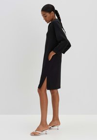 Black shift dress with long sleeves, side pockets, and a side slit. Smooth fabric, minimalist design, mid-thigh length, paired with silver slide sandals.