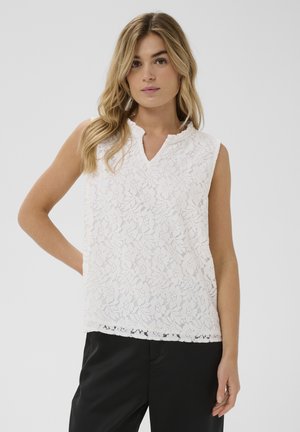 Frau mit langen blonden Haaren, die ein ärmelloses weißes Spitzenoberteil und schwarze Hosen trägt, vor einem schlichten hellgrauen Hintergrund stehend.