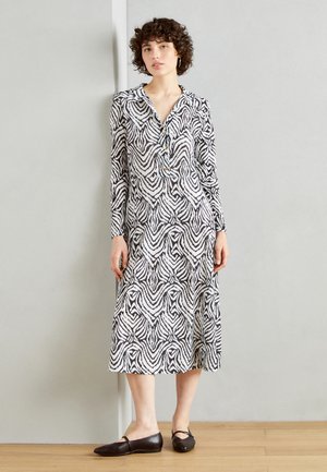 Schwarzes und weißes gemustertes Kleid mit langen Ärmeln, Kragen, Knopfdetails und fließender Silhouette. Getragen mit dunklen flachen Schuhen.