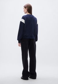 Σκούρο μπλε φούτερ με λευκές λεπτομέρειες στους ώμους, μακριά μανίκια, ριμπ μανσέτες και τελείωμα, σε συνδυασμό με φαρδιά μαύρα παντελόνια, με θέα από πίσω.