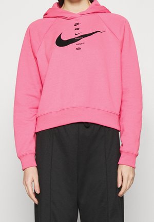 Osoba ubrana w jaskrawy różowy kaptur Nike z czarnymi logo i napisami, w połączeniu z luźnymi czarnymi spodniami na prostym tle.