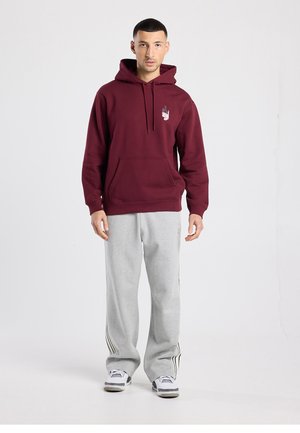 Bordeauxrotes Kapuzen-Sweatshirt mit einer Fronttasche und Grafik; kombiniert mit grauer Jogginghose mit schwarzen Streifen an den Seiten.