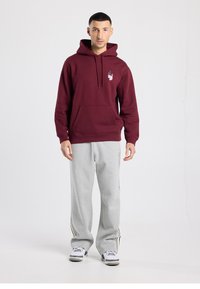 Sweat à capuche bordeaux avec poche avant et motif graphique ; assorti à un pantalon de survêtement gris avec des bandes noires sur les côtés.